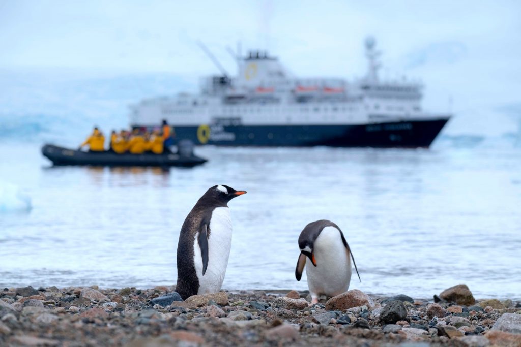 Two penguins in front of a Zodiac boat and a larger boat on the shore in Antarctica.