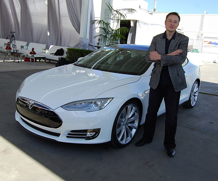 Elon Musk stands in front of a white Tesla. He is reportedly living in Austin.