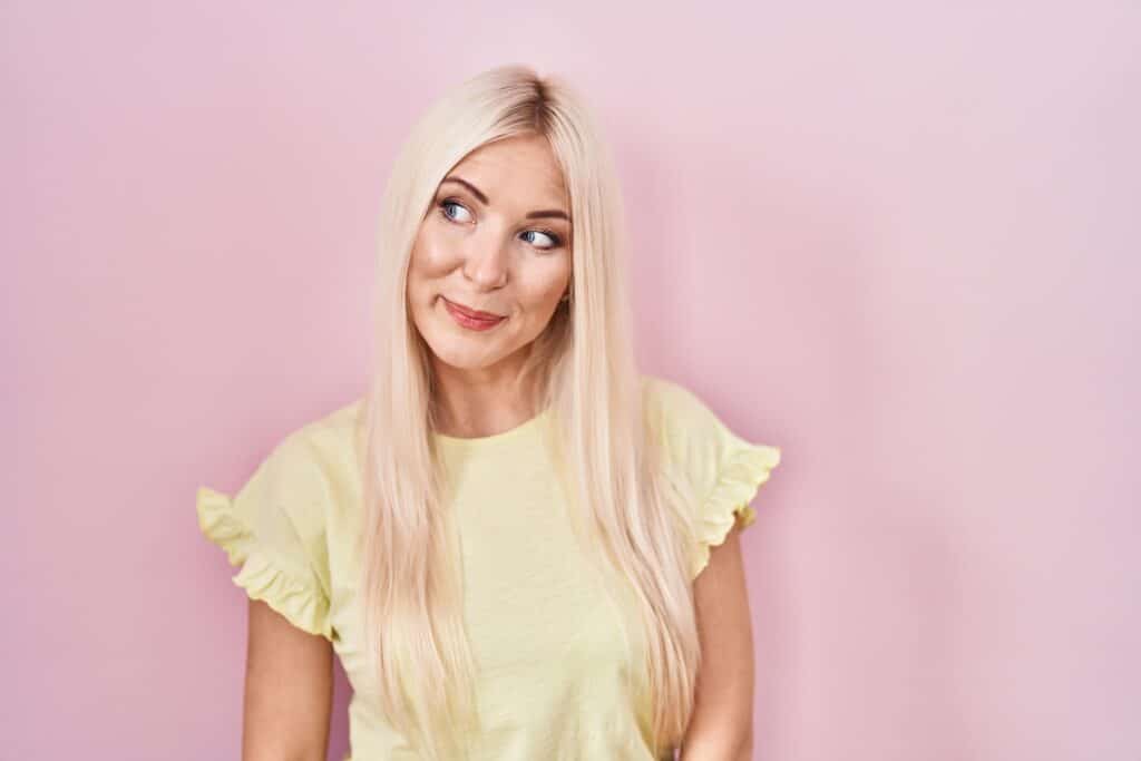 Caucasian woman standing over pink background smiling looking to the side and staring away thinking.