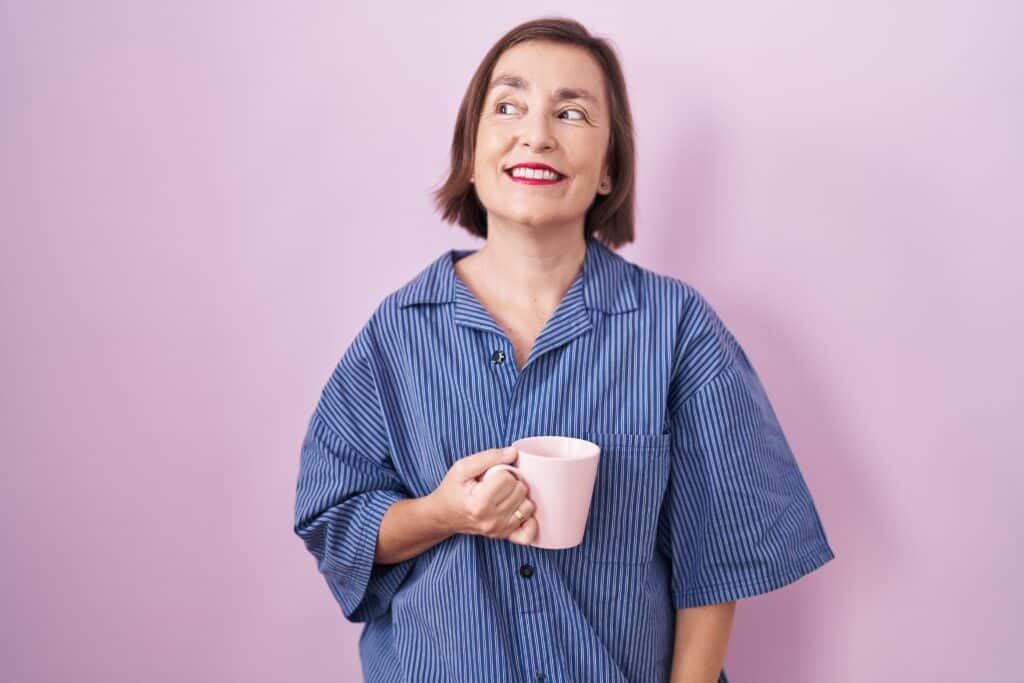 Middle age hispanic woman drinking a cup coffee smiling looking to the side and staring away thinking.